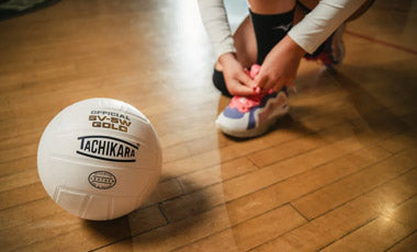 Person tying shoelaces next to a Tachikara volleyball on a wooden floor