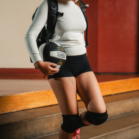 Person holding a volleyball with a backpack, standing on a wooden floor.