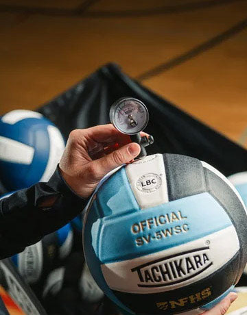Person inflating a tachikara volleyball with a gauge, with additional volleyballs in the background